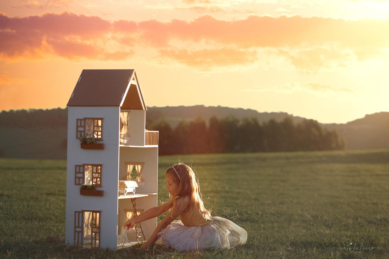 Menina brincando de casinha em um cenário mágico. Campo verde e nuances alaranjadas do sol deixam o ambiente com aparência bucólica.