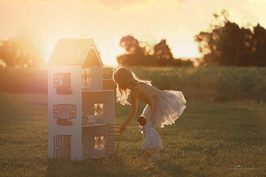Menina brincando de casinha em um cenário mágico. Campo verde e nuances alaranjadas do sol deixam o ambiente com aparência bucólica.