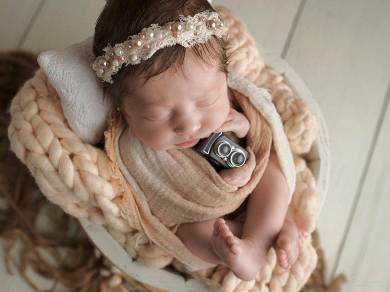 Foto de bebê recém nascido com uma tiara de flores e uma miniatura de câmera nas mãos